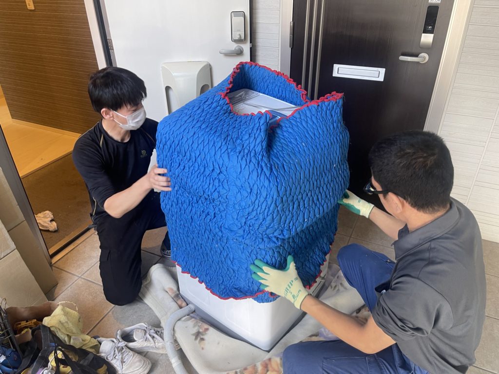 四條畷市の単身引越しで洗濯機を厚手の養生で保護し、室内から安全に搬出するスマイルプロジェクトの作業風景

